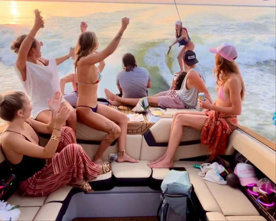 Group of friends on a motorboat at sunset cheering and holding drinks as a wake surfer rides the boat’s wake on open water