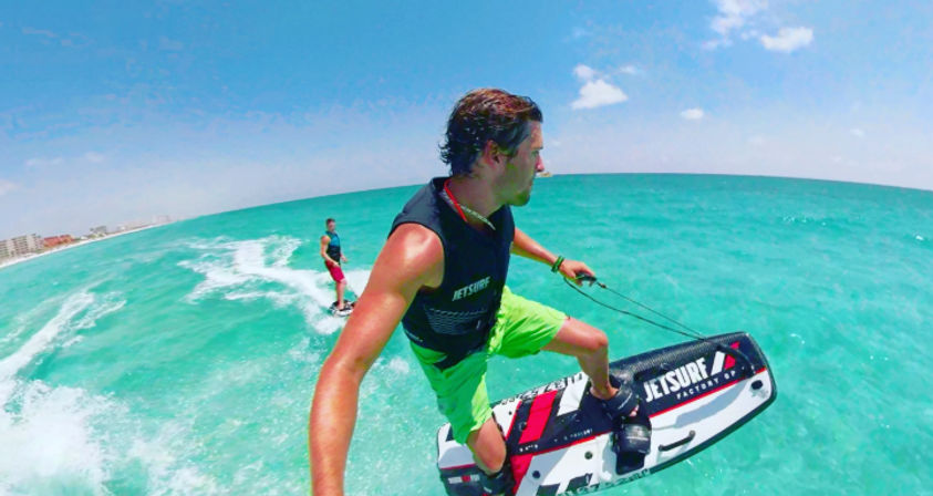 Two riders on powered surfboards skimming bright turquoise coastal waters off a sunny beach; foreground rider in neon green shorts catching air over a small wake.