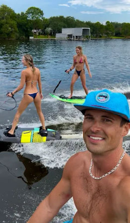 Smiling man in a blue cap takes a lakeside selfie while two women in bikinis wakesurf behind him near a tree-lined shore and boathouse.