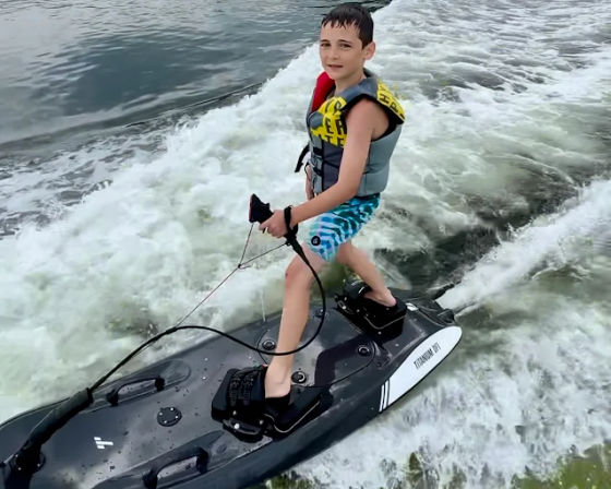 Child in a life jacket standing on a black motorized surfboard, holding a tow handle and carving through a boat wake on a lake — summer watersports scene.