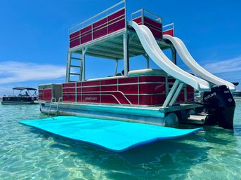 Red double-decker pontoon boat with twin white water slides, outboard motor, and a large blue floating mat in clear turquoise shallow water under a bright blue sky.