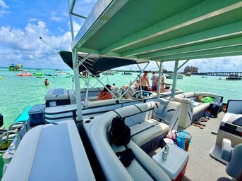 Party-ready pontoon boat with cushioned seating and coolers anchored in clear turquoise water, surrounded by people on inflatables, distant bridge and waterfront buildings under a sunny sky