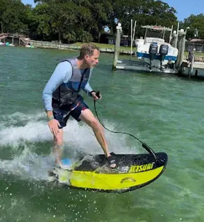 Adult in a life vest shredding a yellow motorized surfboard across green coastal water near a marina dock with a moored boat