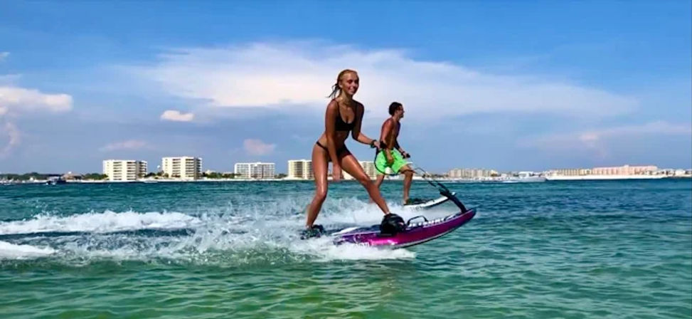 Two people riding electric surfboards across turquoise coastal waters with beachfront condos on the horizon under a sunny blue sky