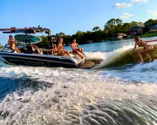 Wakesurfing scene: friends on a wakeboat on a sunny lake with tree-lined shoreline and waterfront houses, a woman riding the boat’s surf wave while others lounge on the stern.