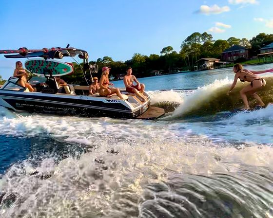Group on a wake-sport boat with a colorful surfboard as a woman wakesurfs behind it on a sunny lake with tree-lined shoreline and waterfront homes