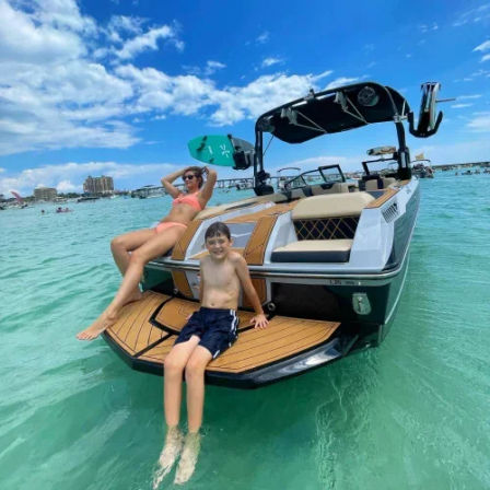 Woman in a pink bikini and a child in swim trunks relaxing on the swim platform of a wakeboard-equipped motorboat in clear turquoise water under a sunny blue sky, beach and boats in the distance.