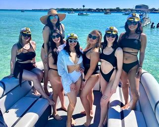 Seven women in swimsuits, sunglasses and captain hats posing cheerfully on a pontoon boat over turquoise water with boats and shoreline in the background.