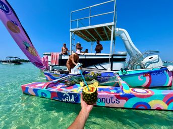 Hand holding a pineapple cocktail toward a colorful painted floating party boat with slide and deck, people socializing on a sunny day in clear turquoise shallow water.