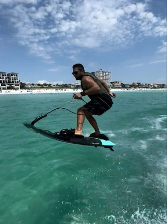 Man riding an electric surfboard (e-foil) above turquoise ocean water near a sandy beach and coastal buildings under a partly cloudy sky.