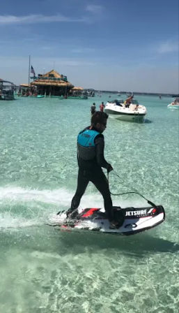 Person riding a motorized surfboard across clear turquoise shallow water near a tiki-style floating bar and anchored boats under a sunny blue sky