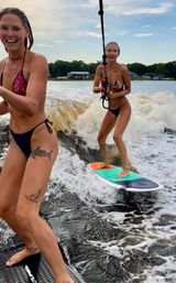 Two women wakesurfing on a summer lake near a tree-lined shore, one smiling while riding a colorful surfboard through the boat wake.