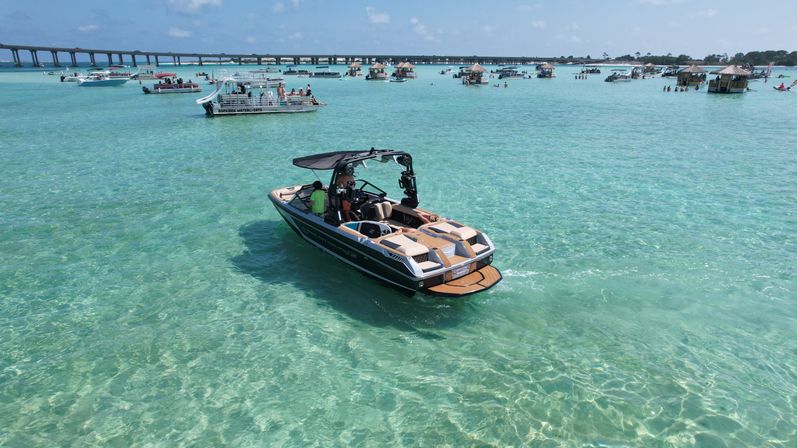 Motorboat cruising over crystal-clear turquoise shallow bay on a sunny day, surrounded by anchored party boats, floating huts and a long coastal bridge in the background.