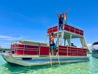 Three people celebrating on a two-level red pontoon boat anchored in shallow, clear turquoise water under a bright blue sky, ladder lowered to the water and other boats in the distance.