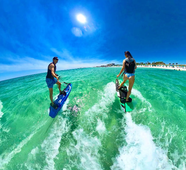 Two riders on electric jetboards gliding over turquoise, crystal-clear ocean waves toward a sunny beach shoreline under a bright blue sky