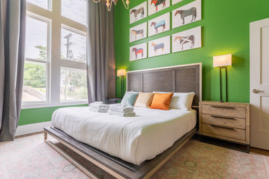 Bright modern bedroom with a bold green accent wall, king bed dressed in crisp white linens and colorful throw pillows, neatly stacked towels on the bed, large floor-to-ceiling windows with gray curtains letting in natural light, wooden headboard and nightstand, playful horse artwork above, and a soft area rug