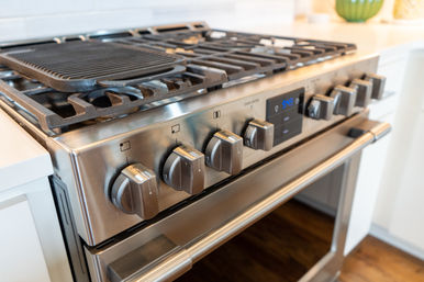 Close-up of a modern stainless-steel gas stove and oven in a bright kitchen, showing a cast-iron griddle on the cooktop, control knobs, and digital display.