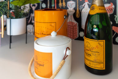 Orange-labeled champagne bottle with matching canister and white ceramic ice bucket with wicker handle on a countertop, potted plant and patterned barware wallpaper in the background.