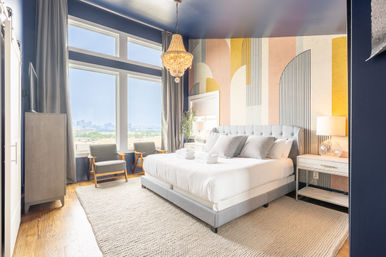 Sunlit modern high-rise bedroom with king bed and tufted gray headboard, pastel geometric accent wall, crystal chandelier, floor-to-ceiling windows framing an urban skyline, two mid-century chairs, bedside table with lamp, and textured area rug.