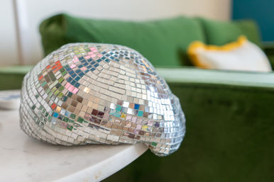 Sparkling mirror-tiled disco helmet on a white marble side table in a living room, reflecting colorful highlights with a green velvet sofa and yellow-trim pillow blurred in the background.