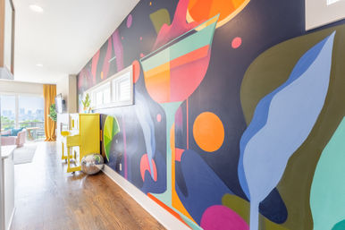 Modern city apartment hallway with hardwood floors, bold colorful mural of an oversized cocktail and abstract shapes, bright yellow piano, disco ball, and windowed living room with skyline view.