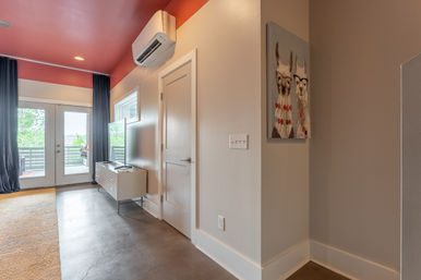 Contemporary urban apartment living area with red ceiling, wall-mounted AC, TV on a white console, polished concrete floor and beige rug, glass doors to a balcony with black curtains, and playful llama artwork on the wall.