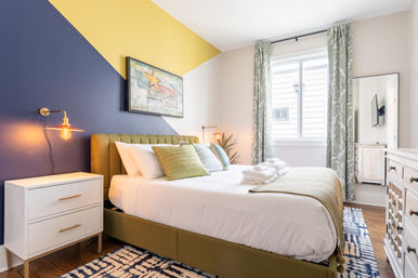 Sunlit modern guest bedroom with green upholstered bed, white linens and folded towels, navy-and-yellow diagonal accent wall, brass wall lamps, white nightstand, patterned rug and curtains, and light wood dresser.