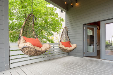 Covered outdoor deck with two hanging wicker egg chairs outfitted with beige cushions and orange pillows, gray siding, leafy green tree and open glass door