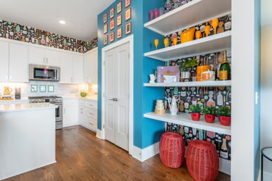 Bright modern kitchen with white cabinets, stainless appliances, teal accent wall and open shelves displaying orange glassware, potted plants and red wicker stools against playful cocktail-pattern wallpaper on warm hardwood floors.