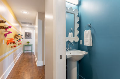 Residential hallway with hardwood floors and colorful high-heel shoe display on a yellow-striped wall leading to a small blue powder room with white pedestal sink, scalloped mirror, and towel ring