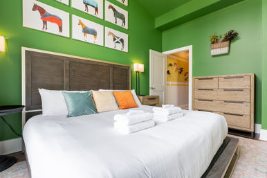 Bright green guest bedroom with king bed, crisp white linens and folded towels, colorful throw pillows, rustic wood headboard and dresser, horse-themed wall art and open doorway to a sunny yellow hallway.