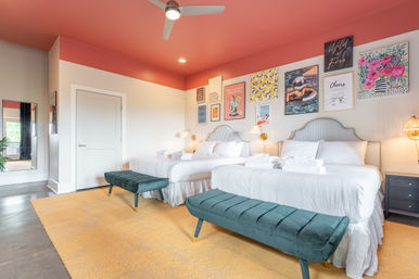 Bright modern guest bedroom with two white-dressed beds, coral-pink ceiling, colorful gallery wall, teal velvet benches, yellow rug and ceiling fan