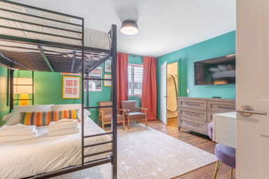 Cheerful teal-walled guest room in a vacation rental with a metal bunk bed over a made double bed with striped cushion and folded towels, wooden dresser with wall-mounted flat-screen TV, two mid-century armchairs, coral curtains and hardwood floors.