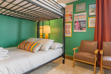 Cozy modern guest room with a black metal bunk bed, white bedding and a colorful striped pillow against a green accent wall, framed pop-art posters, a warm floor lamp, two brown armchairs and red curtains.