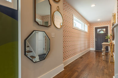 Stylish residential entry hallway with chevron-pattern wallpaper, hardwood floors, three decorative wall mirrors, narrow clerestory windows, dark front door and small console table