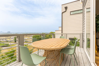 Rooftop balcony with oval wooden dining table and sage-green chairs on a gray wood deck, cable railing overlooking a highway and distant city skyline under a cloudy sky.
