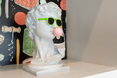 White classical bust statue wearing neon green sunglasses and blowing a pink bubblegum bubble, displayed on a white shelf against a colorful patterned backdrop as quirky modern decor.