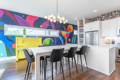 Bright modern kitchen with white island and six black bar stools, colorful abstract wall mural, yellow upright piano, globe chandelier, stainless steel refrigerator and hardwood floors
