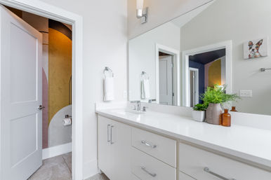 Bright modern bathroom with white vanity and long countertop, undermount sink and chrome faucet, large frameless mirror, potted plant and amber bottles on the counter; open door reveals colorful geometric accent wall and toilet.