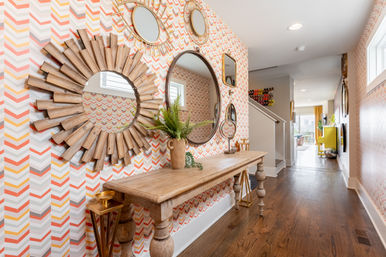 Sunlit modern hallway entry with playful chevron orange-yellow-gray wallpaper, rustic wood console table, sunburst and round mirrors, hardwood floors and open living area beyond