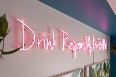 Playful pink neon script sign reading 'Drink Responsibly' mounted on a pale wall above framed botanical prints in a modern interior with a teal ceiling and hanging plants.