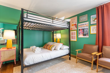 Cheerful green bedroom with a black metal bunk bed, neatly made lower bed with striped accent pillows and folded white towels, mid-century wooden armchairs, coral curtains, bedside lamps, and colorful framed wall art