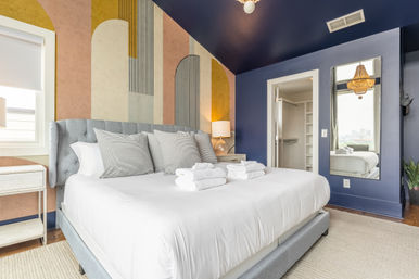 Sleek urban bedroom with a plush king bed, colorful geometric accent wall and deep navy ceiling, mirror reflecting a city view, bedside lamp and neatly folded towels