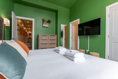Cheerful green bedroom with white linens and neatly folded towels on the bed, colorful throw pillows, wall-mounted flat-screen TV, wooden dresser and mirror, and an open doorway revealing a hallway with decorative wall-mounted accents.