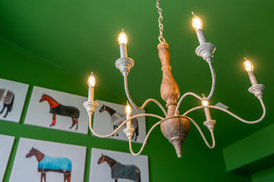 Rustic wooden chandelier with six lit bulbs hanging from a chain against a vivid green ceiling, framed colorful horse prints displayed on the wall below
