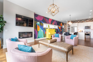 Bright open-plan living room with pastel pink armchairs and teal pillows, tufted leather bench, bold abstract wall mural and a yellow upright piano beside a white modern kitchen with stainless appliances.