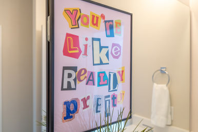 Framed colorful bathroom wall art with ransom-note style letters on a pink background reading You're Like Really Pretty, reflected in a modern vanity mirror with towel ring and small plant