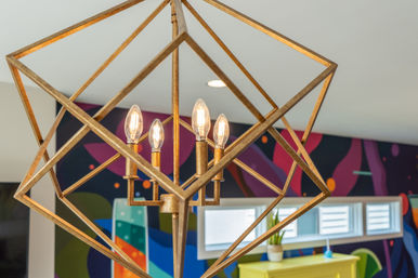 Gold geometric pendant chandelier with five exposed candle-style bulbs in a bright contemporary interior featuring a colorful abstract mural, narrow windows, and a yellow console table.