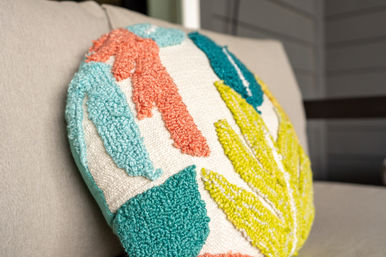 Round tufted throw pillow with teal, coral and lime abstract leaf pattern resting on a beige sofa — textured modern home decor accent.
