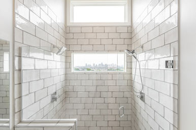 Bright, sunlit modern shower with white subway tiles, glass door, built-in bench, dual rain and handheld showerheads, and a narrow window framing a distant city skyline.